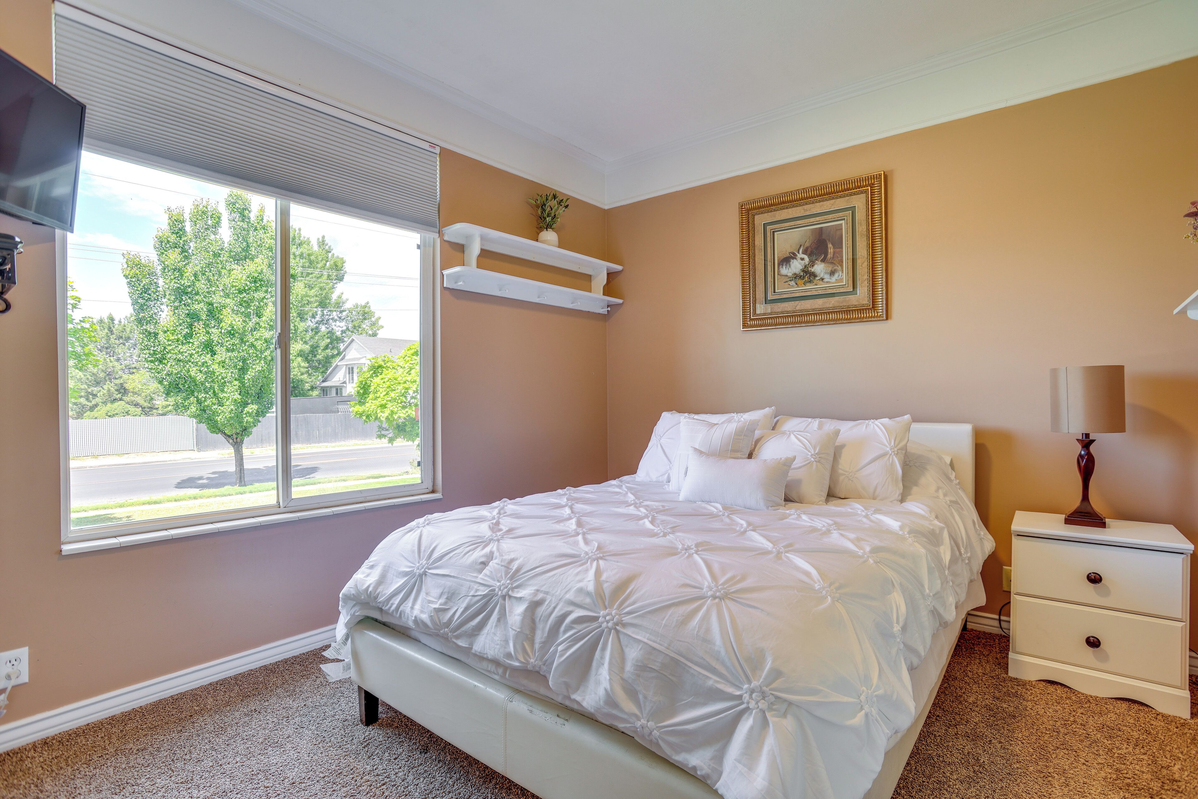 A bedroom with a bed, a nightstand, a TV, a window with blinds, and a wall-mounted shelf.