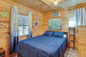 A wooden cabin room with a bed, a painting, a shelf, and a window with curtains. - Smoky Mtn Tiny Cabin Hideaway: Hot Tub, Patio (Englewood)