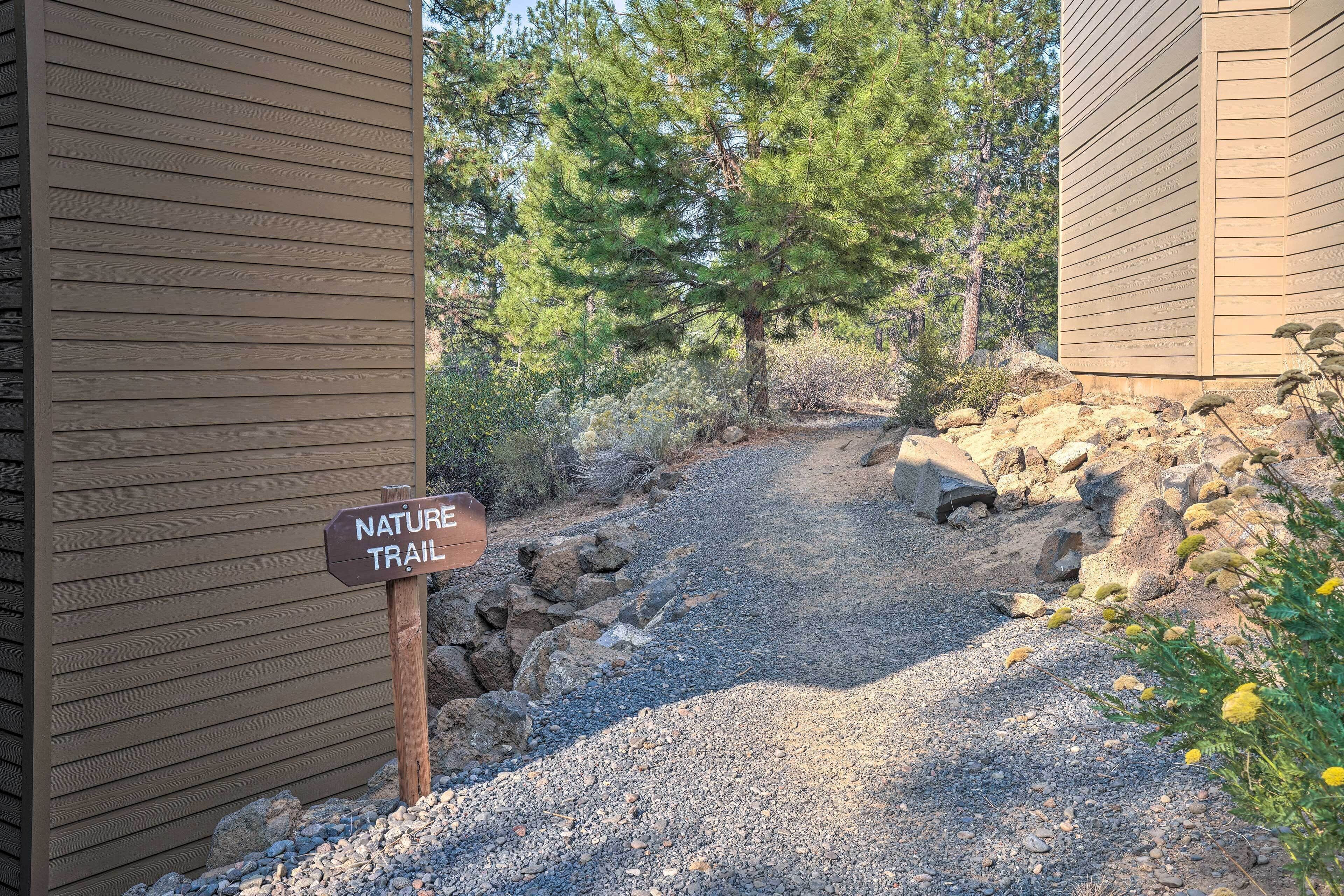 Un sentier de gravier mène à un panneau indiquant le début d’un sentier de découverte de la nature, le tout étant entouré d’arbres et de rochers.