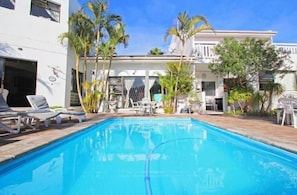 A swimming pool with lounge chairs, palm trees, and a white building with multiple windows and balconies.