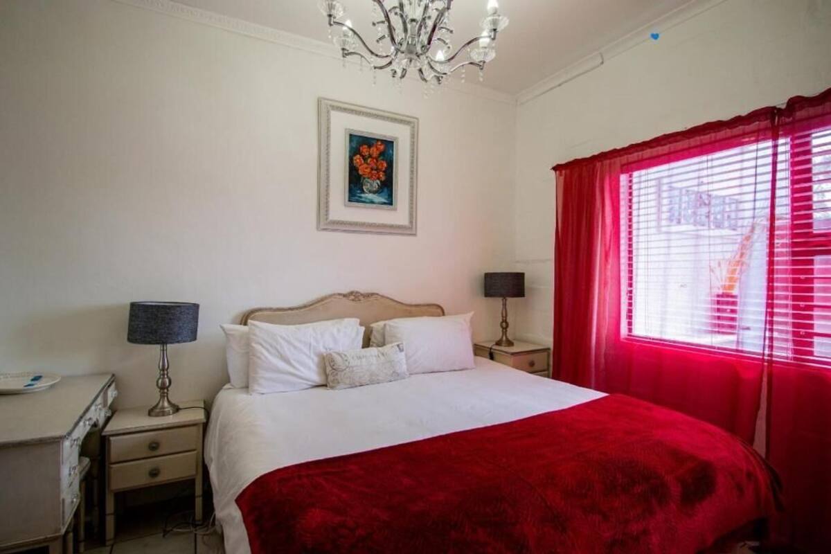 Un dormitorio con una cama, dos mesitas de noche, una lámpara, un cuadro enmarcado en la pared y una ventana con cortinas rojas.