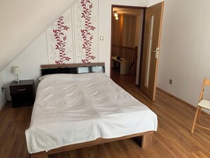 Um quarto com cama, mesa de cabeceira, abajur, cadeira de madeira e porta que dá para uma escada.