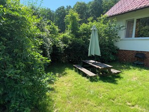 A backyard with a picnic table and an umbrella, surrounded by dense greenery and a house with a red roof.