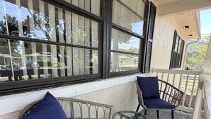Un balcon avec des fauteuils en osier et une table en verre, de grandes fenêtres avec des volets, et une vue sur le quartier.