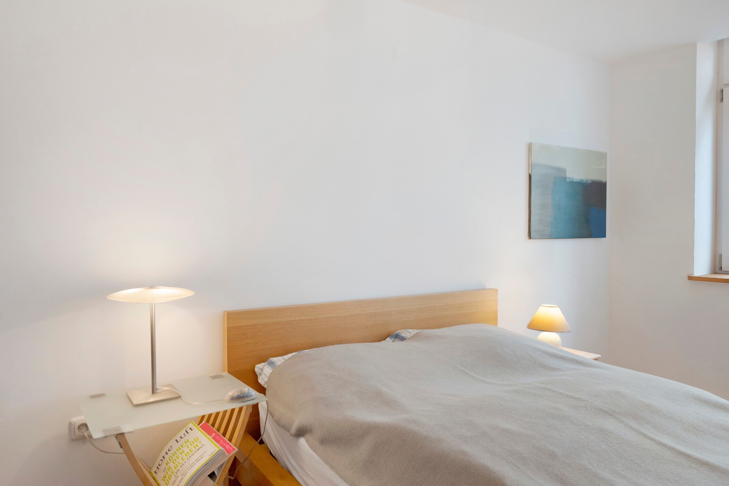 A bedroom with a bed, bedside lamps, a nightstand, a wall-mounted painting, and a book on the nightstand.
