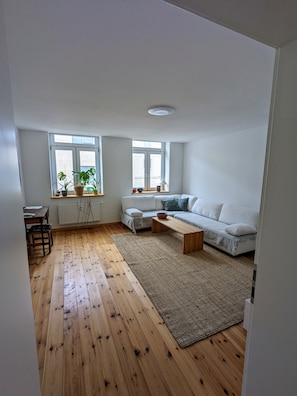 A modern living room with a wooden floor, a white sofa, a coffee table, and a dining area with a table and chairs. - Apartment in Schwerin (Schwerin)