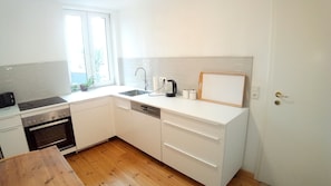 A compact kitchen with a dishwasher, stove, and a window with a plant. - Apartment in Schwerin (Schwerin)