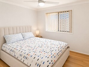 Une chambre à coucher comprenant un lit, une table de chevet, une lampe, un ventilateur de plafond et une fenêtre avec des stores.