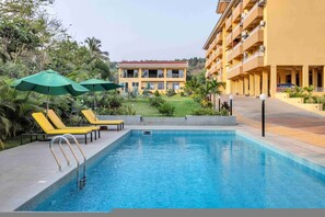 Ein Pool mit Liegestühlen und Sonnenschirmen, umgeben von Grünflächen und einem Gebäude mit Balkonen.