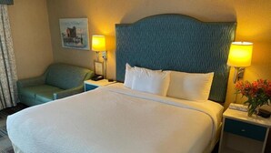 Una habitación de hotel con una cama grande, dos lámparas de noche, una mesita de noche, una silla y un cuadro en la pared.