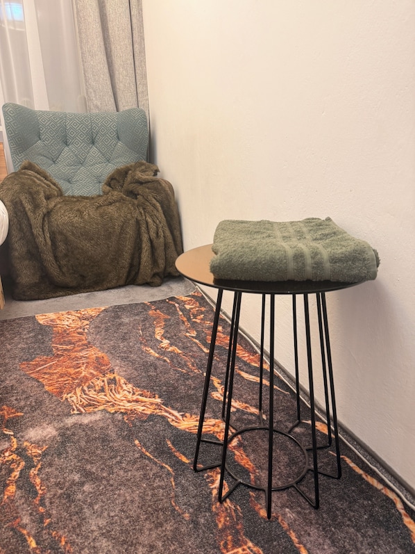 A corner of a room with a patterned rug, a small stool, a chair with a blanket, and a wall with a curtain.