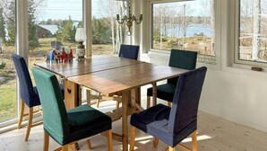 Een eethoek met een houten tafel en stoelen, grote ramen met uitzicht op bomen en een meer, en een kroonluchter.