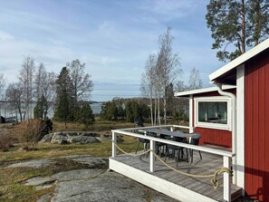 A wooden deck with outdoor furniture, a view of a lake, and a red building with a window. - Property with sauna and jetty near Grisslehamn (Grisslehamn)