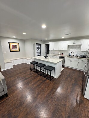 A modern kitchen with white cabinets, a central island with stools, a built-in washer and dryer, and a grey couch in the living area. - Charming 3BR Atlanta Bungalow • Minutes to Midtown & Mercedes‑Benz Stadium (Atlanta)