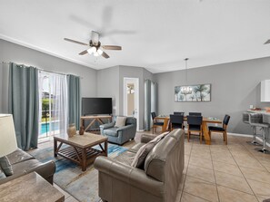 Ein modernes Wohnzimmer mit Sofa, Fernseher, Esstisch mit Stühlen und Deckenventilator.