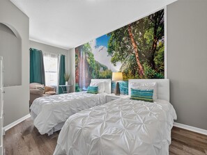 Ein Hotelzimmer mit zwei Betten, einem Nachttisch, einer Lampe, einem Fenster mit Vorhängen und einem Wandbild von einem Wasserfall und Wald.