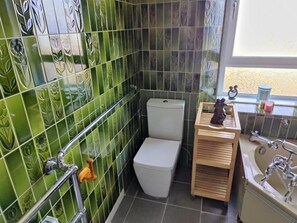 Ein Badezimmer mit grün gemusterten Fliesen, einer weißen Toilette, einem hölzernen Waschtisch mit Spiegel und einer Badewanne.