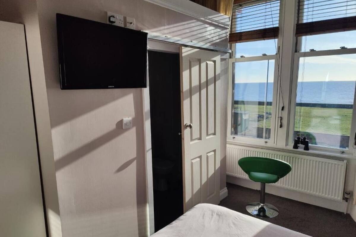 A room with a bed, a television mounted on the wall, a window with a view of the sea, and a radiator.