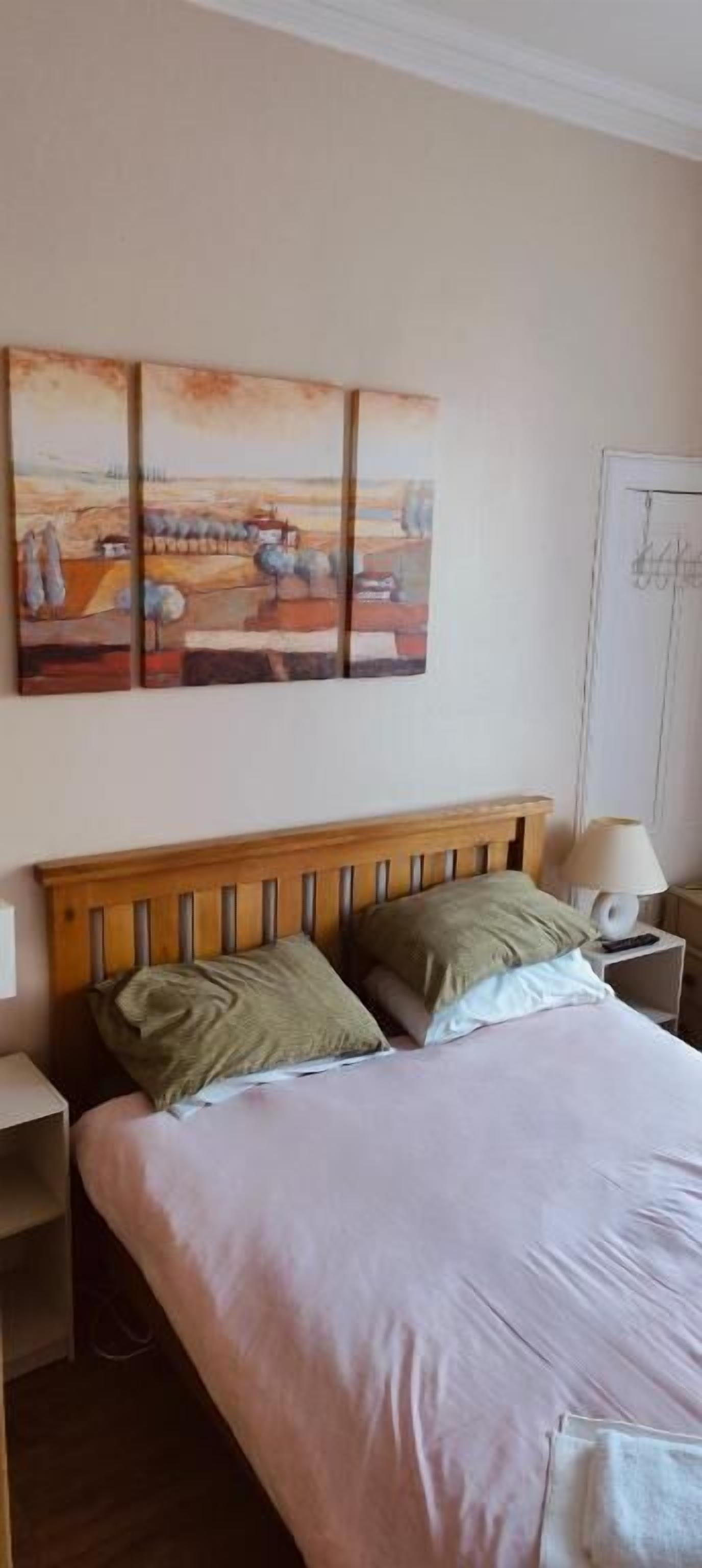 A bedroom with a wooden headboard, a bed with two pillows, a nightstand with a lamp, and a wall-mounted painting.