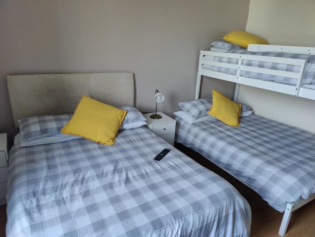A room with two beds, each with a checkered bedspread and a single yellow pillow. There is a nightstand with a lamp between the beds.