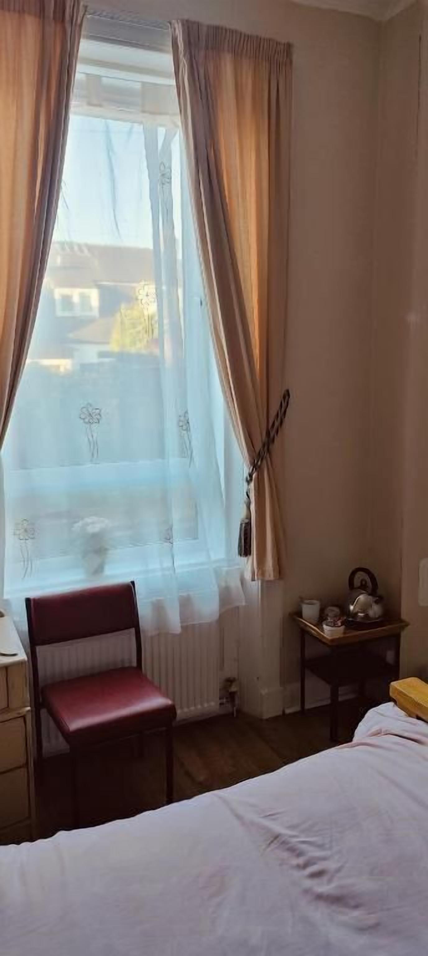 Ein Zimmer mit Bett, Sessel, Tisch, Fenster mit Vorhängen und Heizkörper.
