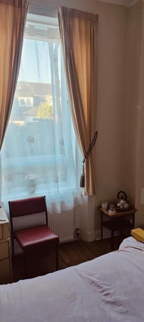Ein Zimmer mit Bett, Sessel, Tisch, Fenster mit Vorhängen und Heizkörper.