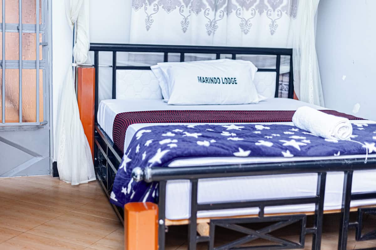 Sängyssä on valkoinen tyyny, jossa lukee teksti »MARINDO LODGE», sininen kuviollinen peitto ja puinen pääty.