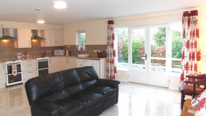 A living room with a black leather sofa, a dining area with a table and chairs, a kitchen with white appliances, and a sliding glass door leading to a balcony. - Ballyvoy Camping Barn (Ballycastle)
