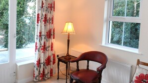 A room with a chair, a table with a lamp, a window with a view of trees, and a radiator. - Ballyvoy Camping Barn (Ballycastle)