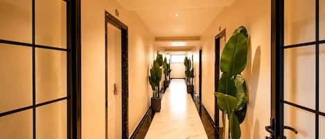 Un pasillo de hotel con piso de mármol, plantas en macetas y puertas con marcos negros.