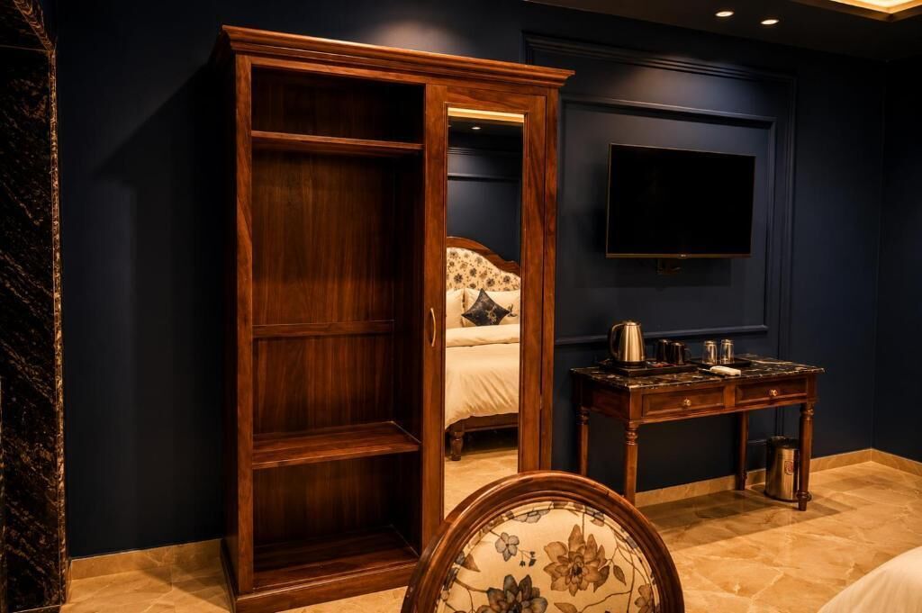 A room with a wooden wardrobe, a bed with a floral-patterned headboard, a small table with a kettle, and a TV mounted on the wall.