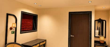 A room with a wooden door, a mirror, a small table, a chair, and a ceiling light.