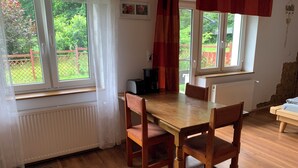 Un coin repas avec une table et des chaises en bois, un radiateur, une fenêtre donnant sur un espace verdoyant et un rideau rouge.