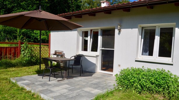 Un espace patio avec une table et des chaises, un parasol marron et un bâtiment blanc avec un toit rouge.