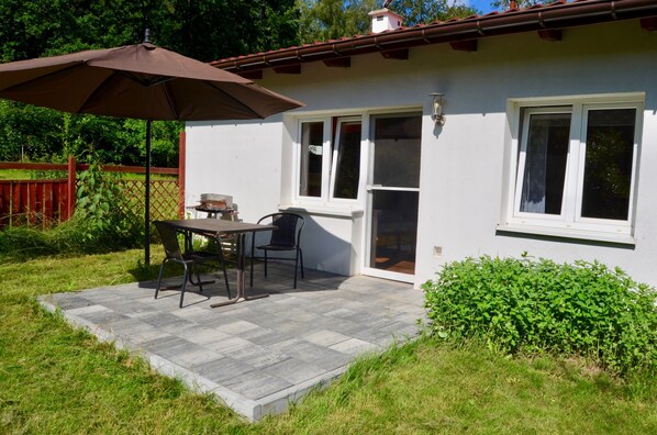 A patio area with a table and chairs, a brown umbrella, and a white building with a red roof.