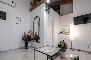 Ein Zimmer mit Fliesenboden, einem Tisch mit Spitzenbettdecke, einer Topfpflanze, einem Wandspiegel und einem Flachbildfernseher.