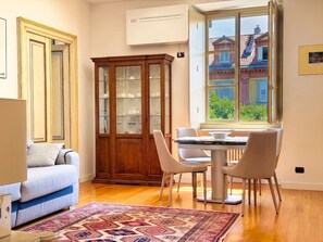 Ein Zimmer mit Esstisch, Stühlen, einer Couch, einem Schrank und einem Fenster mit Blick auf Gebäude.