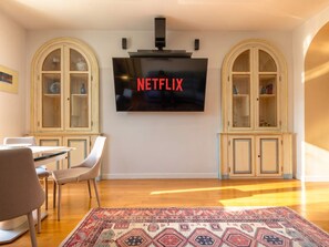 Ein Wohnzimmer mit einem an der Wand montierten Flachbildfernseher, einer Essecke mit Stühlen und einem Tisch sowie zwei Bogenunterschränken mit Glastüren.