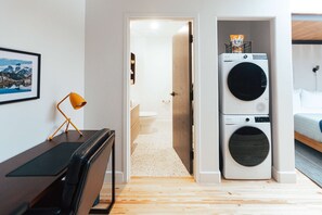 A room with a desk, chair, two stacked washing machines, a bed, and a door leading to a bathroom.