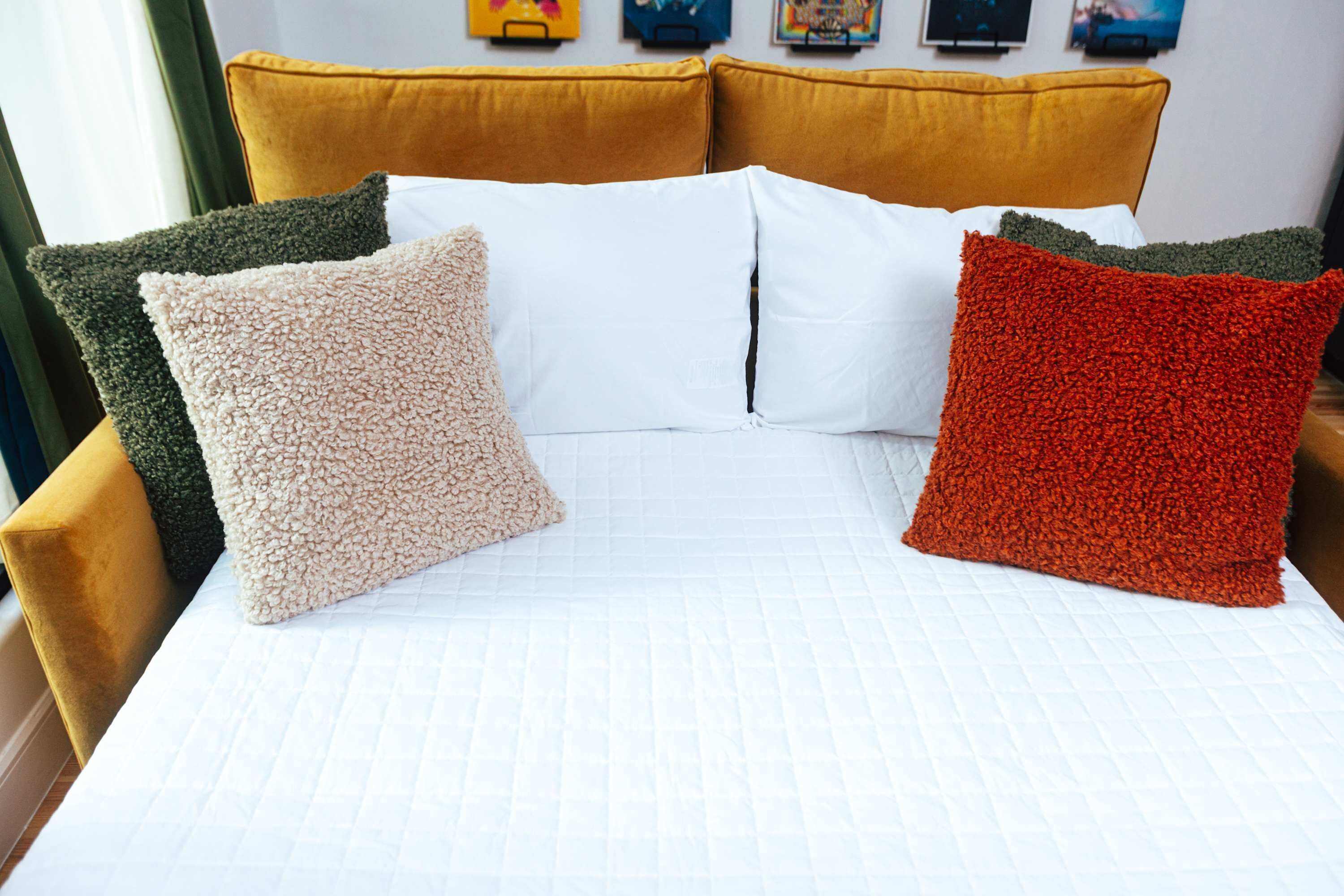 Una cama con colcha blanca acolchada, cabecera color mostaza y cuatro almohadas de varios colores.