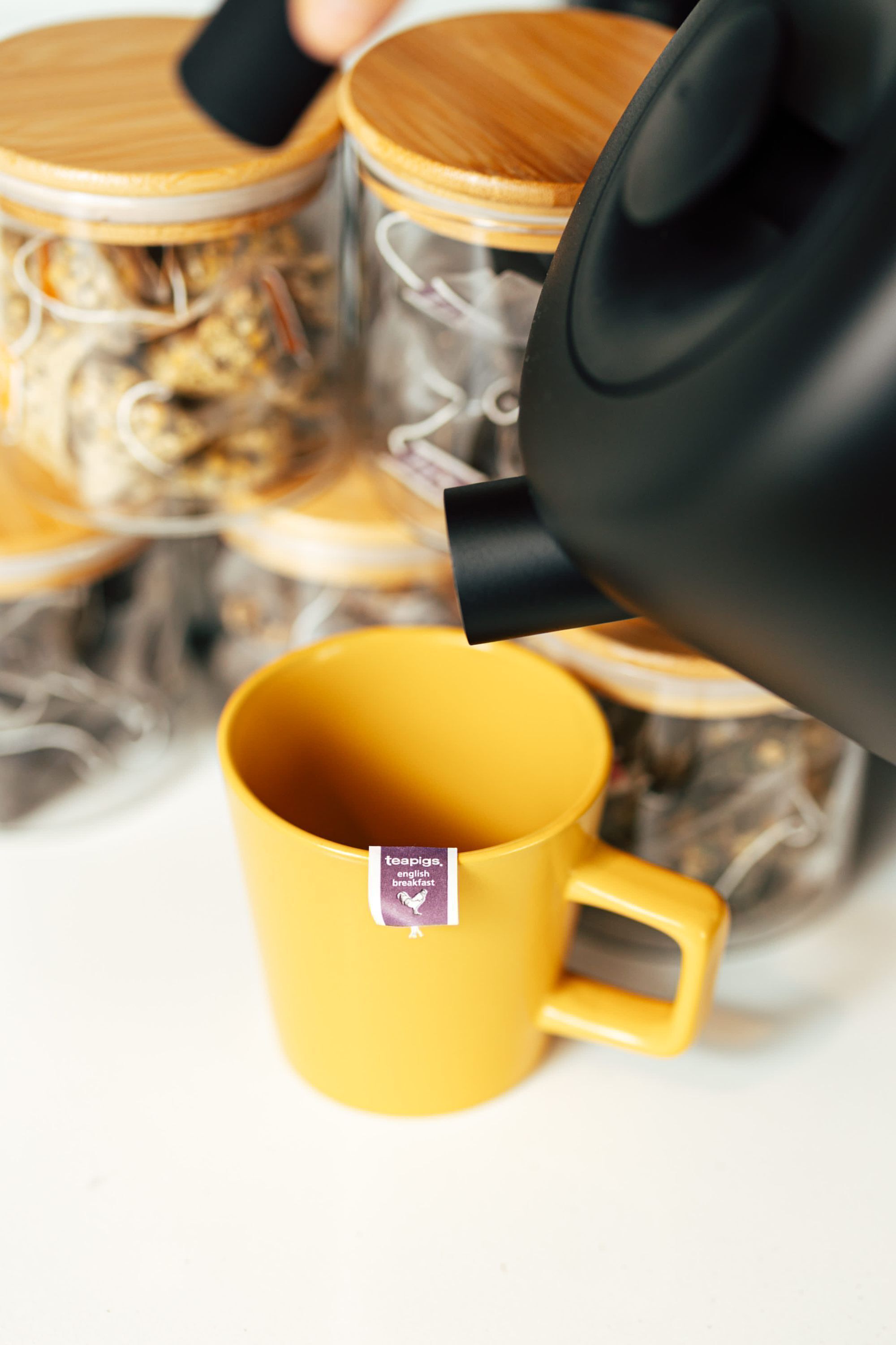 Una taza amarilla con etiqueta de bolsita de té, un hervidor negro vertiendo en ella, y un fondo desenfocado con un objeto de madera.