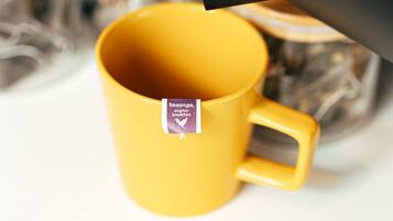 Una taza amarilla con etiqueta de bolsita de té, un hervidor negro vertiendo en ella, y un fondo desenfocado con un objeto de madera.