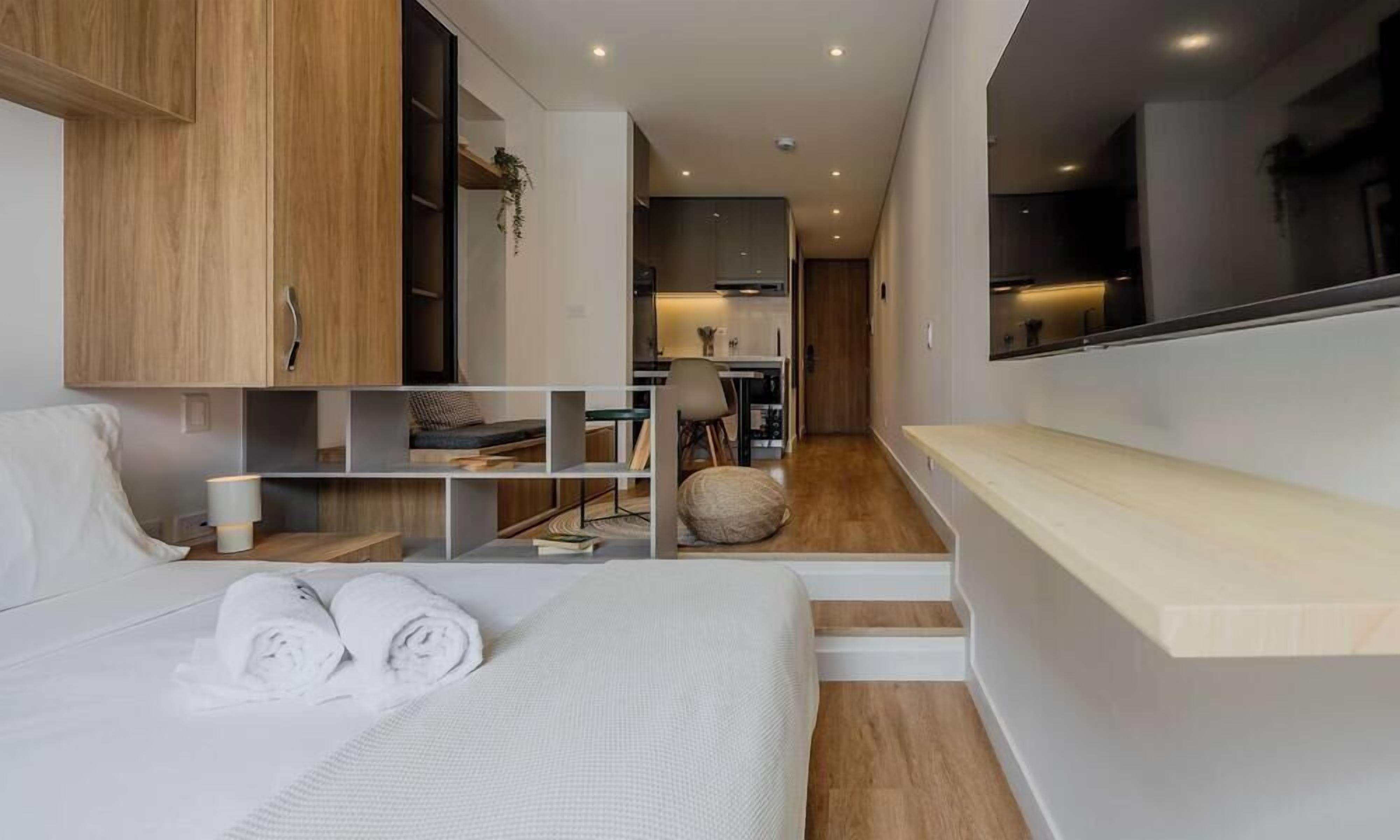 A modern hotel room with a bed, a desk, a chair, and a small kitchen area.