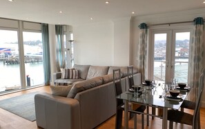 A modern living room with a sofa, a dining table set for four, and large windows overlooking a marina. - Spinnaker Apartment 3 bed Waterside Apartment (Cowes)