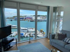 Ein modernes Wohnzimmer mit großem Fenster mit Blick auf eine Marina, einem Flachbildfernseher und einem grauen Sofa.