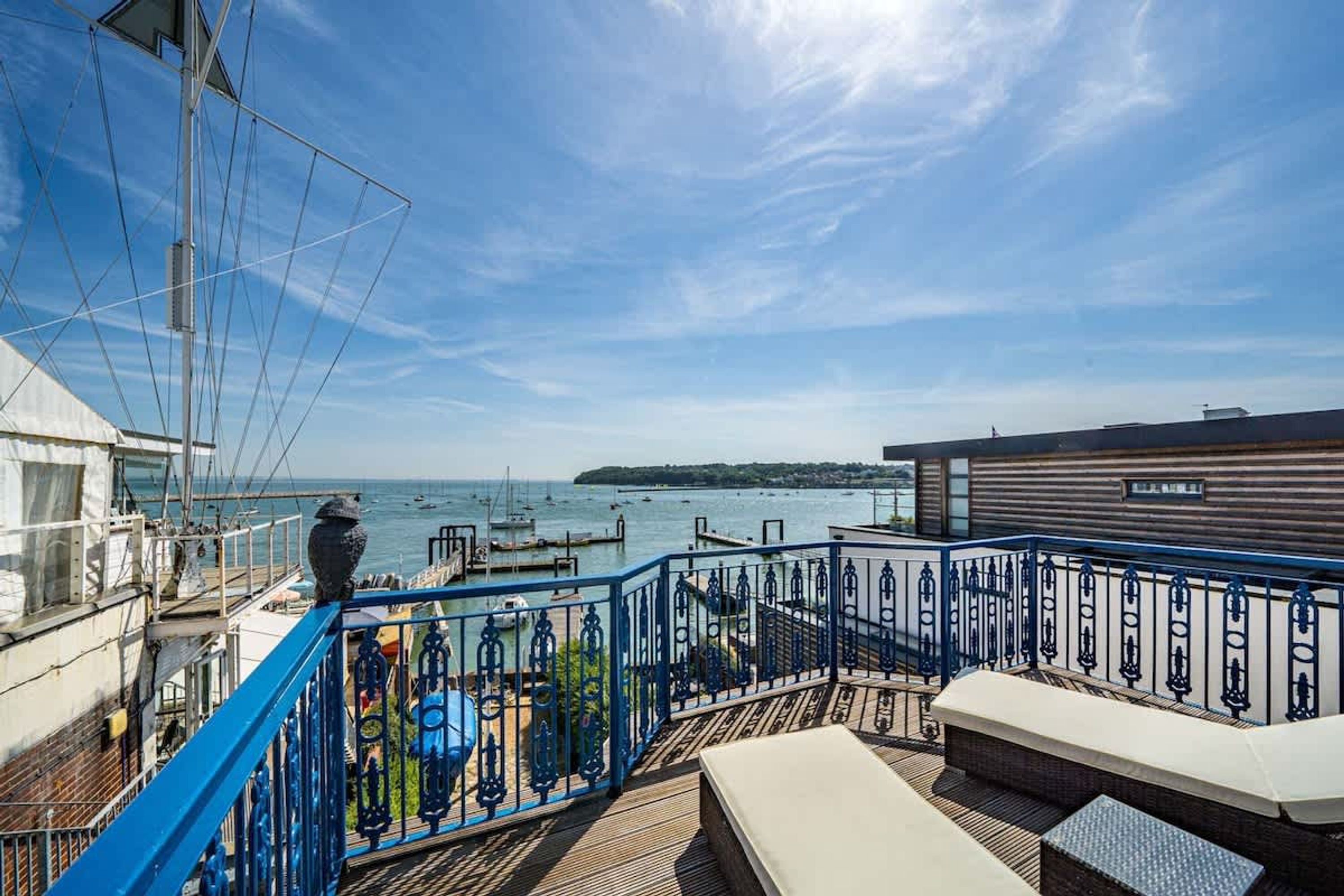 Une terrasse dotée d’une rambarde bleue, d’un banc blanc et offrant une vue sur la mer et les bateaux.