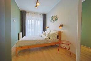 Quarto com cama de madeira, uma mesinha vermelha e janela com cortinas.