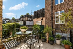 Une terrasse avec une table et des chaises en verre, entourée de plantes en pot et d’un mur en briques.