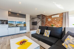Un salon moderne avec un canapé noir, une table basse blanche ornée de fruits, une cuisine en arrière-plan et un mur de briques avec une horloge.