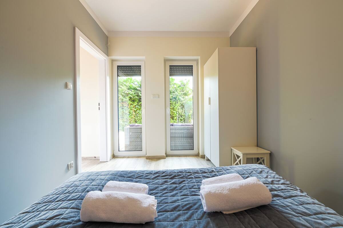 Un dormitorio con una cama, dos toallas blancas, una mesita pequeña y vistas a zonas verdes a través de una puerta.